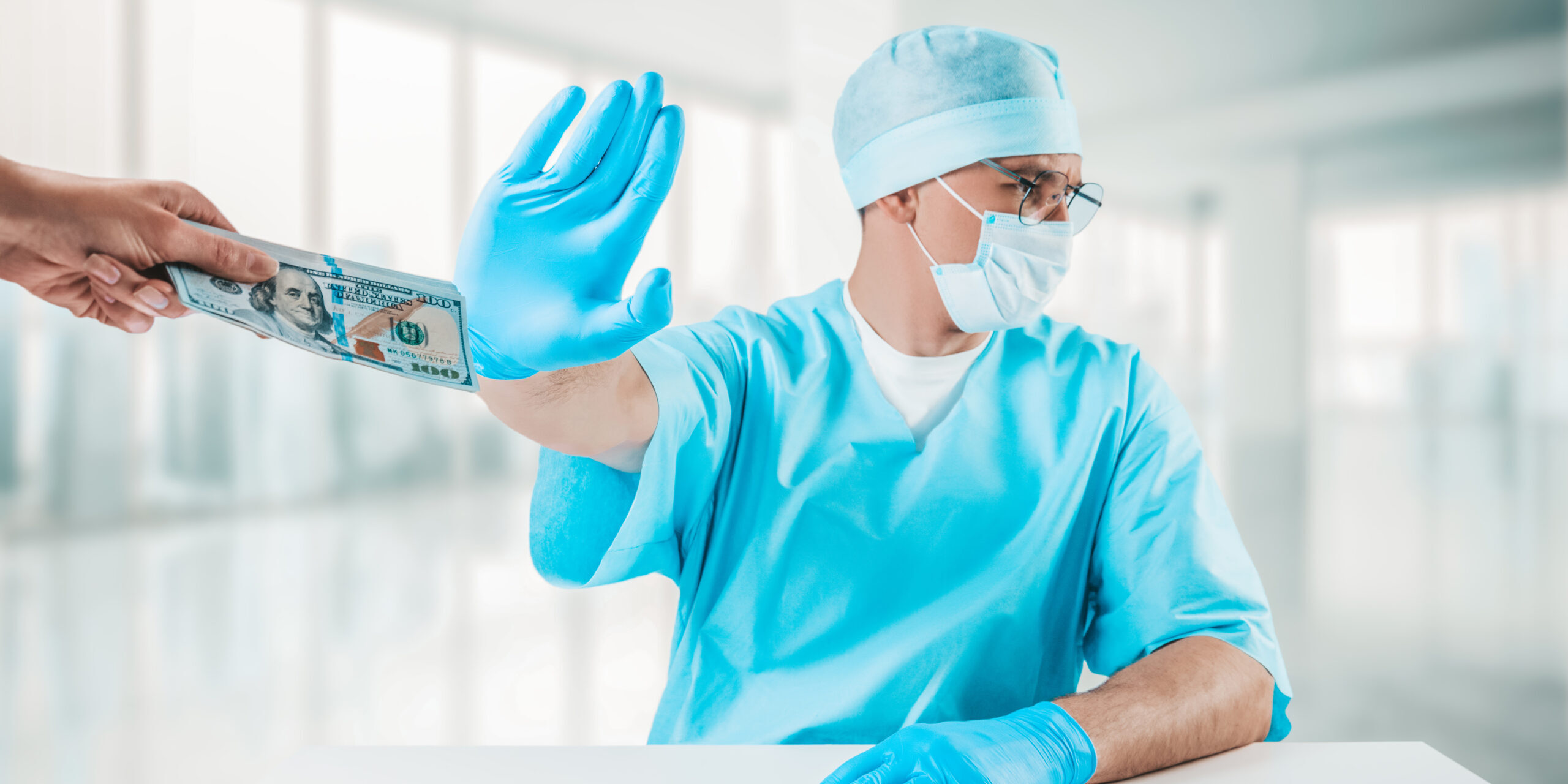 Male doctor in scrubs holds up his hand to refuse cash being handed to him | Article on Stark Law enforcement | Vanguard Communications | Denver