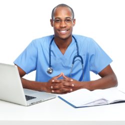 African-American doctor seated at a table with a stethoscope around his neck.  He is wearing blue scrubs and is sitting in front of a laptop and open book.