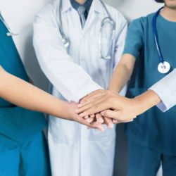 A doctor with a white coat standing between two nurses with blue scrubs, all three are in a huddle with their hands out signifying they are a happy team working together