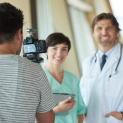 Photo of a male doctor in a white coat next to a female medical practitioner in blue scrubs standing in front of a person with a camera on a tripod