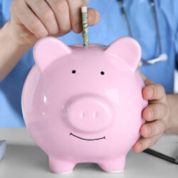Physician putting bills into a pink piggy bank, avoiding one of the common mistakes in meeting financial goals | Vanguard Communications | Denver, CO