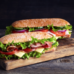 Two fresh submarine sandwiches with ham, cheese, bacon, tomatoes, lettuce, cucumbers and onions on wooden cutting board in a blog explaining how to use the sandwich technique for employee engagement feedback | Vanguard Communications | Denver, CO