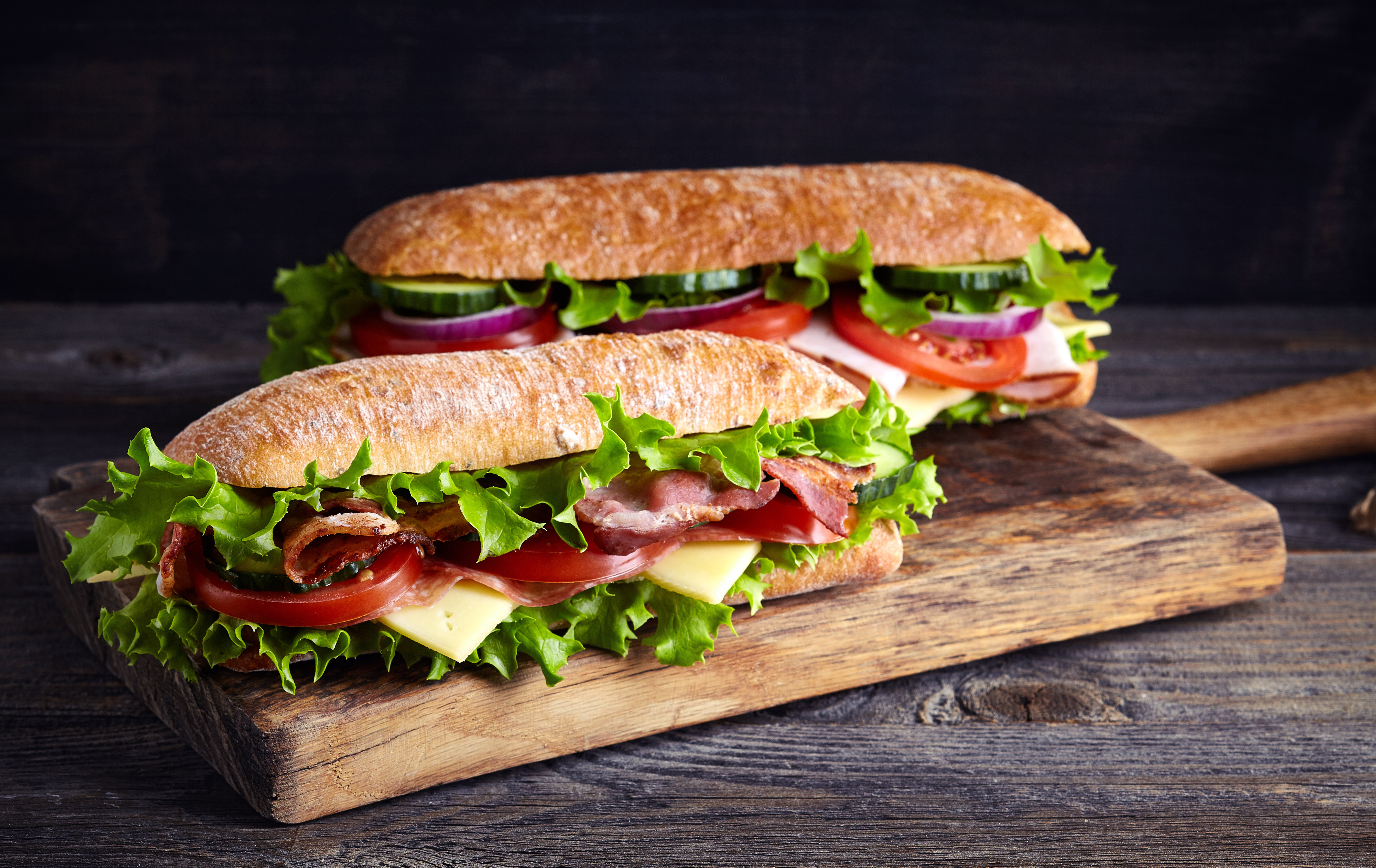 Two fresh submarine sandwiches with ham, cheese, bacon, tomatoes, lettuce, cucumbers and onions on wooden cutting board in a blog explaining how to use the sandwich technique for employee engagement feedback | Vanguard Communications | Denver, CO